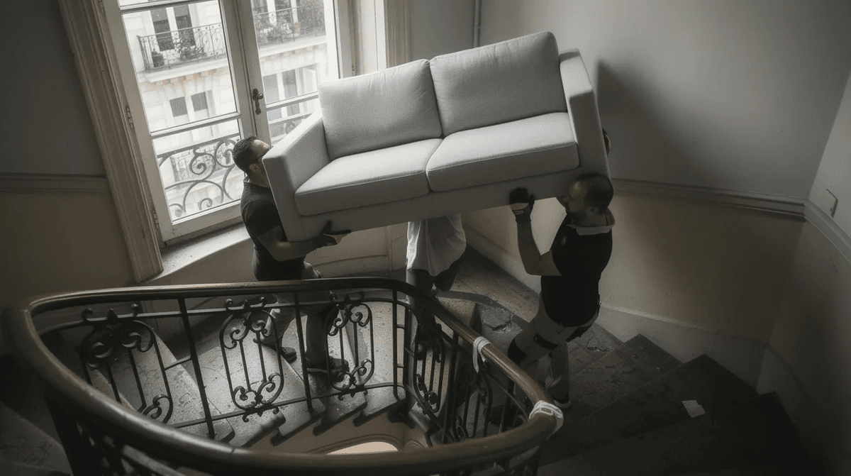 Une équipe de livreurs transporte un canapé à travers un escalier étroit d'un immeuble parisien, illustrant le défi de la livraison de mobilier dans un environnement urbain. Cette scène met en avant le service de livraison clé en main pour les clients souhaitant meubler leur logement avec des équipements de qualité.