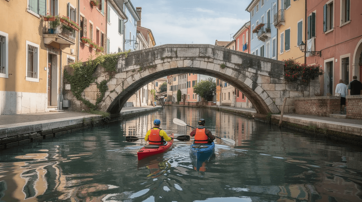 Deux kayakistes pagaient sous un vieux pont en pierre dans un canal d'une ville européenne, entourés de bâtiments colorés sur les deux rives. Cette scène évoque une belle activité de canoë kayak, parfaite pour une balade en famille au cœur de la ville.