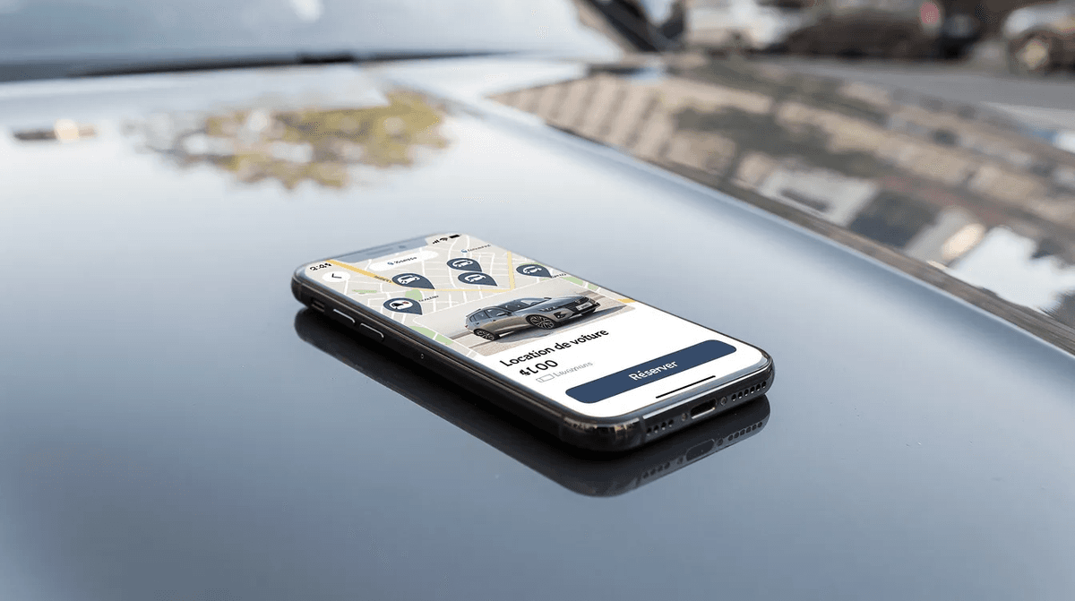 L'image montre un smartphone posé sur le capot d'une voiture, affichant une application de location de véhicules. On peut voir l'interface de l'application qui permet aux particuliers de réserver des voitures pour leurs séjours, avec des informations sur les tarifs et les lieux disponibles.