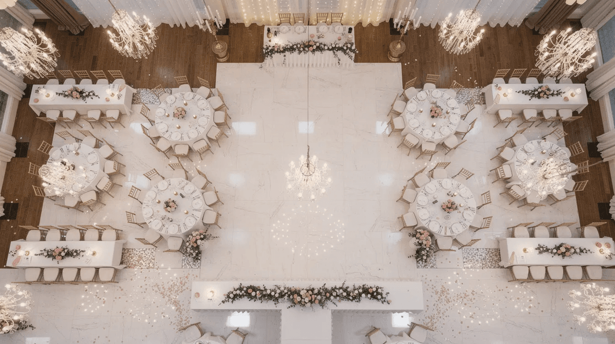 Une vue aérienne d'une salle de réception élégamment aménagée pour un mariage, avec des tables rondes et rectangulaires disposées harmonieusement. L'ambiance festive est accentuée par des décorations raffinées, créant un espace accueillant pour les convives.