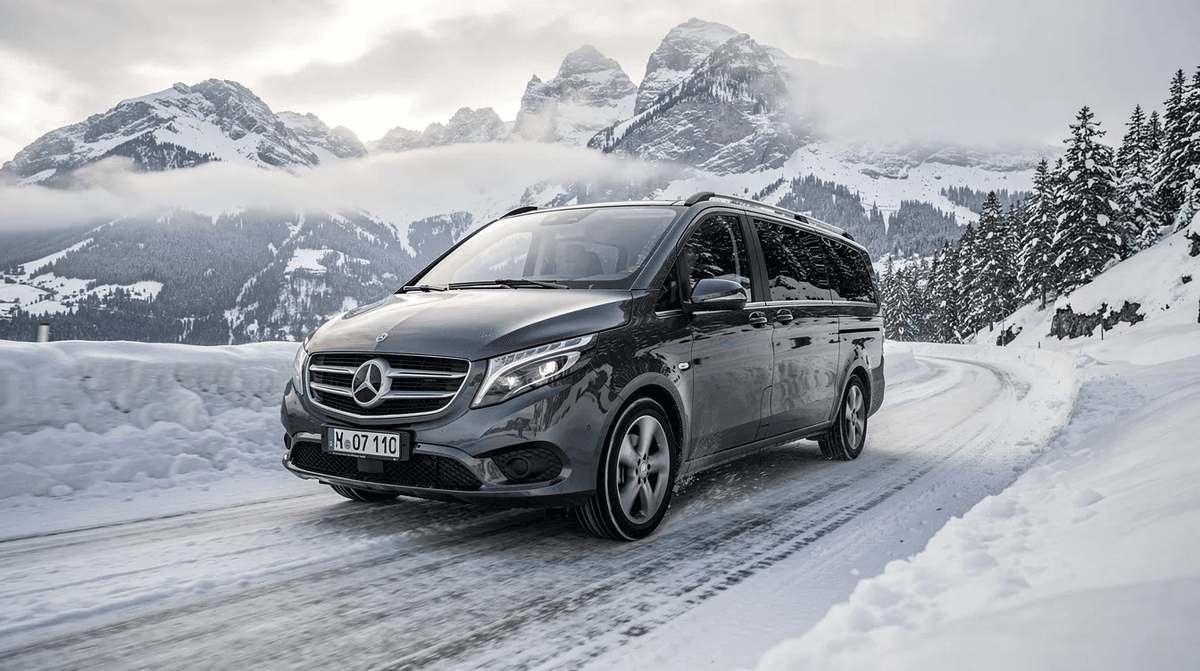 Une Mercedes Classe V haut de gamme roule sur une route de montagne enneigée, avec des sommets alpins majestueux en arrière-plan. Ce minibus, conçu pour le confort des familles et des voyageurs, offre une expérience de conduite agréable dans un paysage hivernal.