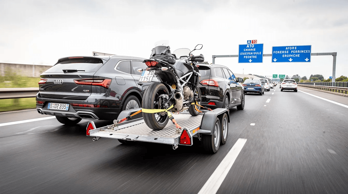 Une moto est solidement fixée sur une remorque porte moto, tractée par un SUV sur une autoroute française. L'image montre le véhicule en mouvement, mettant en avant la sécurité du transport grâce à des sangles et une rampe de chargement.