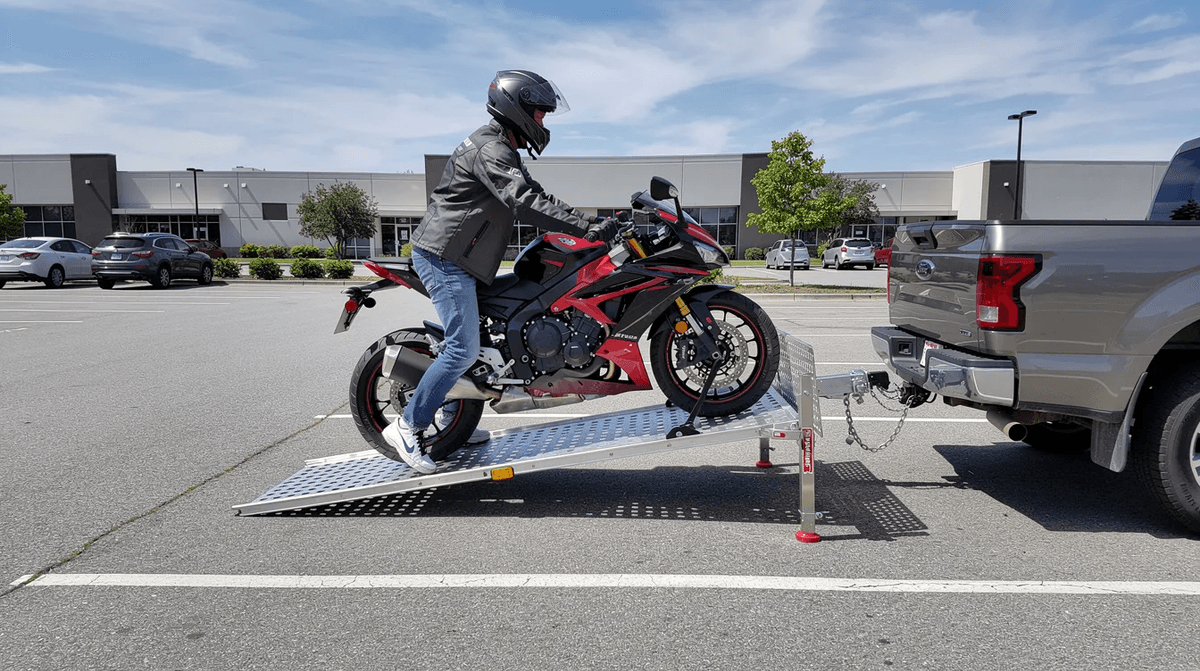 Une personne charge une moto sportive sur une remorque porte moto à l'aide d'une rampe en métal, dans un parking. On peut voir des sangles prêtes à sécuriser le véhicule pour le transport.